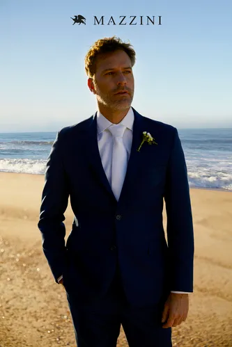 Foto de campaña de Mazzini de un hombre en una playa con traje azul marino, corbata blanca y flor en el ojal